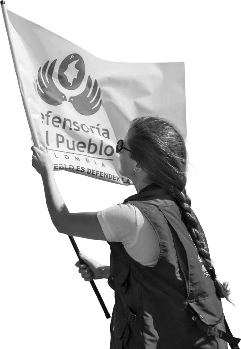Una mujer de espaldas, con chaleco y trenza, ondea la bandera de la Defensoría del Pueblo de Colombia con el lema «Defender al pueblo es defender la vida».
