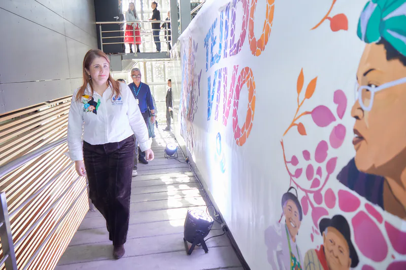 Una funcionaria de la Defensoría del Pueblo camina por una rampa junto al mural de la campaña Tejido Vivo, con el retrato en acuarela de una mujer con turbante.