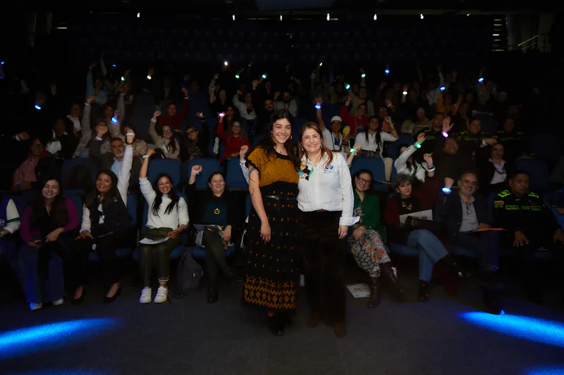 Dos mujeres posan de pie en el auditorio mientras el público, en pie tras ellas, levanta pulseras luminosas.