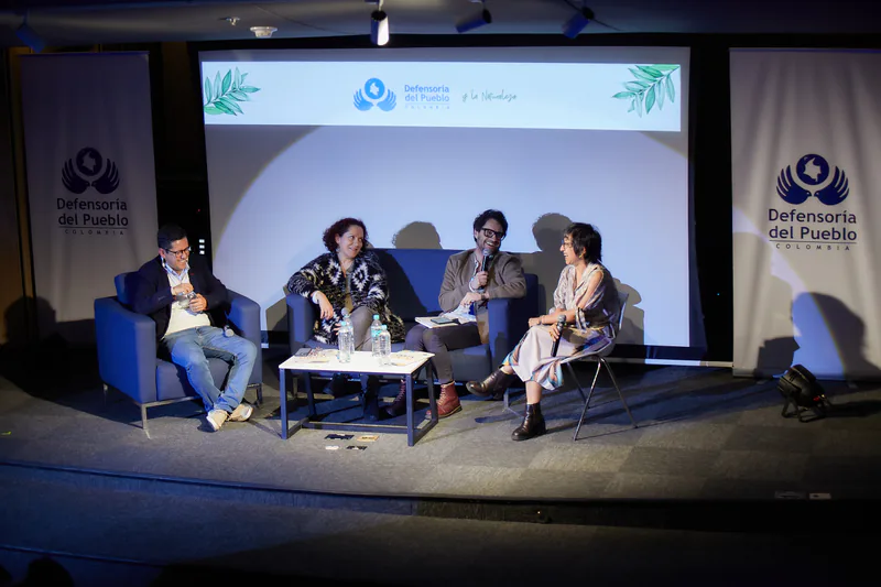 Panel de cuatro personas conversando en el escenario, ante la pantalla con el logotipo de la Defensoría del Pueblo y la Naturaleza.